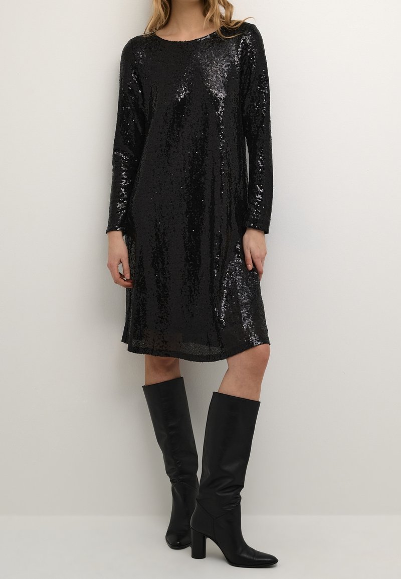 Kvinna som bär en svart långärmad paljettklänning och svarta knähöga klackstövlar, stående mot en enfärgad ljus vägg.