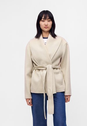 Femme aux cheveux noirs raides portant un manteau beige ceinturé par-dessus un jean bleu sur un fond blanc uni.