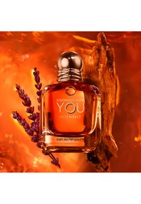 Klar glassparfume flaske med et rundt sølvhett, inneholdende en oransje væske og hvit tekst. Aksentuert med lavendel- og tre-elementer på en livlig bakgrunn.