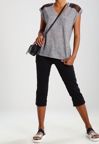 Nainen, joka on pukeutunut harmaaseen lyhythihaiseen paitaan, jossa on mustat verkkokankaiset olkapaneelit, mustiin capri-housuihin, hopeisiin ja tummansinisiin sandaaleihin, ja kantaa mustaa olkalaukkua.