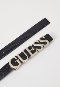Czarny pasek z sztucznej skóry z błyszczącą złotą klamrą z logo "GUESS". Pasek ma teksturowane wykończenie i klasyczny design.