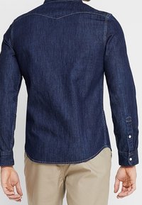 Ciemnoniebieska koszula dżinsowa z długimi rękawami, spiczastym kołnierzykiem i zaokrąglonym dołem. Posiada wzór na yoke i zapinane na guziki mankiety. Gładka tekstura.