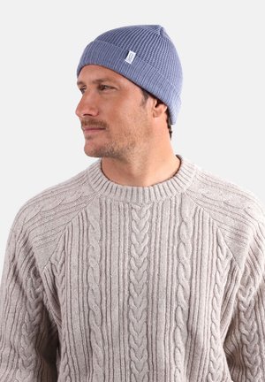 Bonnet tricoté en bleu avec des côtes, doté d'un revers et d'une petite étiquette en tissu blanc. Porté avec un pull en maille torsadée gris clair.