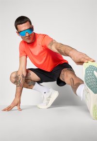 Neon orange træningsskjorte, sorte shorts, hvide sneakers med grønne detaljer, synlige tatoveringer og blå spejlbriller; strækposition.