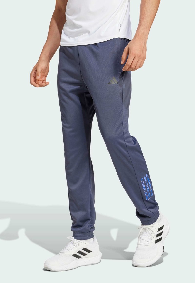 Pantalones deportivos de un azul oscuro con una textura suave, que presentan un logo de Adidas y detalles en azul. Combinados con zapatillas blancas y una camiseta blanca.