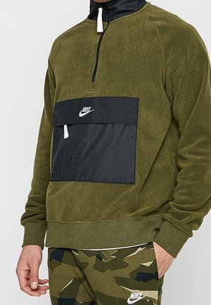 Mężczyzna w oliwkowo-zielonym pulowerze Nike z czarną kieszenią na piersi i dopasowanych moro spodniach Nike, stoi z jedną ręką w kieszeni.