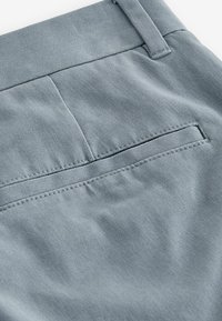Pantaloni de bumbac gri deschis, cu un buzunar plat la spate cu un finisaj curat și o textură subtilă. Detaliu de talie ajustată vizibil.
