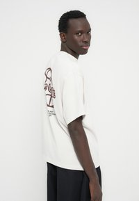 Φωτεινό μπεζ T-shirt με κοντά μανίκια, με μεγάλο μαρουν από γραφικό στο πίσω μέρος και κείμενο. Κανονική εφαρμογή και απαλό βαμβακερό ύφασμα.