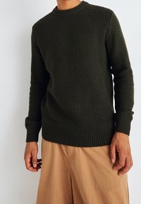 Mörkgrön stickad tröja med ett strukturerat mönster, rund halsringning och ribbade manschetter och nederkant, i kombination med beige byxor.