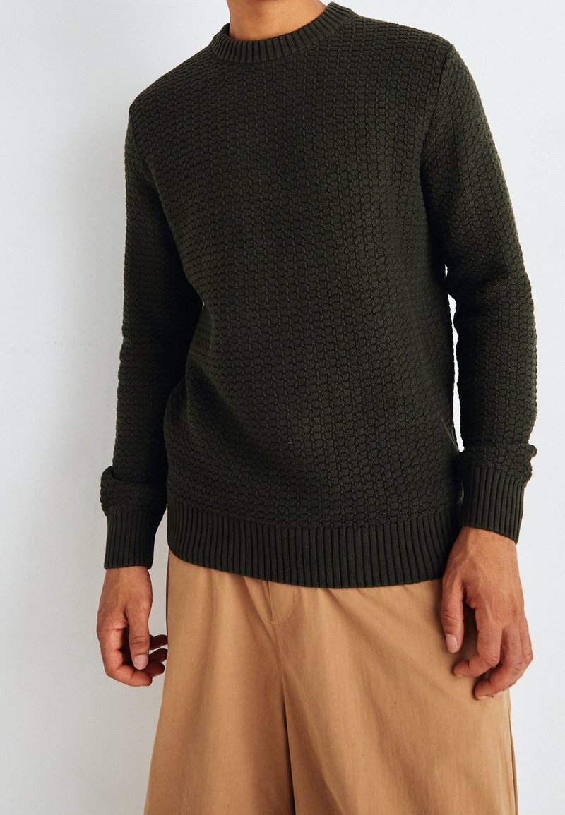 Mörkgrön stickad tröja med ett strukturerat mönster, rund halsringning och ribbade manschetter och nederkant, i kombination med beige byxor.