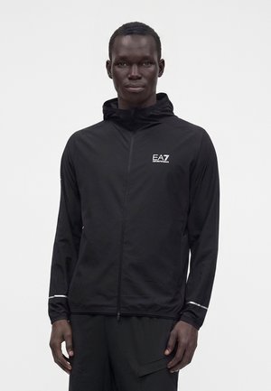 Mužský model nosí černou zipovou bundu EA7 Emporio Armani s kapucí a reflexními pruhy na rukávech, stojící na bílém pozadí.