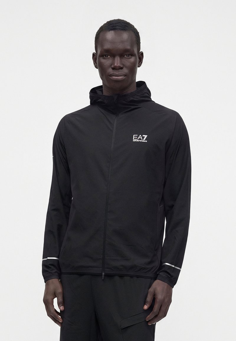 Άνδρας μοντέλο φοράει μαύρο μπουφάν EA7 Emporio Armani με φερμουάρ και κουκούλα, με ανακλαστικές ρίγες στα μανίκια, στέκεται μπροστά σε λευκό φόντο.