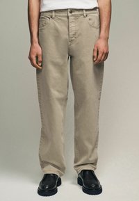 Beige cotton straight-leg trousers with front pockets, belt loops, and a button closure, paired with black leather shoes.