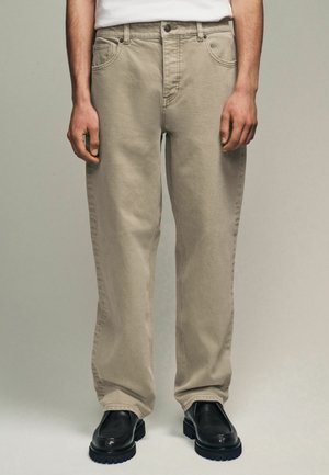 Beige cotton straight-leg trousers with front pockets, belt loops, and a button closure, paired with black leather shoes.