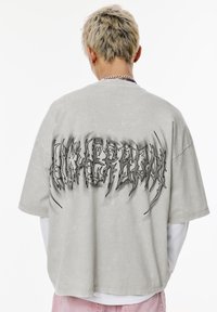 T-shirt in cotone grigio oversize con una finitura testurizzata. Presenta un'audace e frastagliata grafica nera sul retro, maniche corte e una vestibilità comoda.