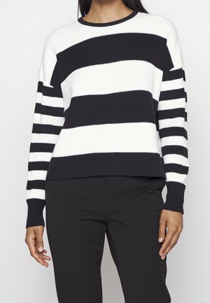 Ein Erwachsener trägt einen schwarz-weiß horizontal gestreiften Pullover mit langen Ärmeln und schwarze Hose, steht vor einem einfarbigen Hintergrund.