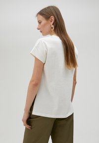 Camisa de algodón blanca con un corte relajado y mangas enrolladas, combinada con pantalones verdes oliva. Textura lisa, diseño sencillo y sin patrones visibles.