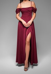 Femme portant une robe de soirée bordeaux épaules dénudées avec une fente haute à la cuisse et des talons ouverts à perles, debout devant un fond gris.