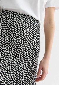 Jupe noire avec un motif carré abstrait blanc. Fabriquée dans un tissu à texture lisse. Design ajusté visible à la taille, associée à un haut blanc.