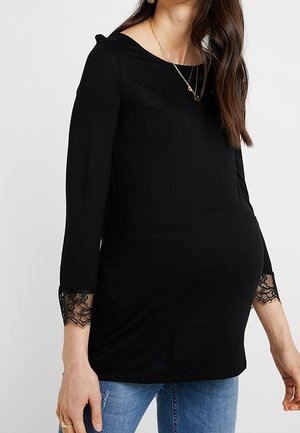 Kvinde iført en tætsiddende sort langærmet top med blonderkanter og blå jeans, der viser en gravid mave, med lagdelte halskæder.