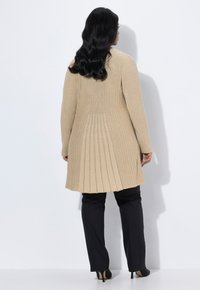 Mujer con largo cabello negro ondulado vestida con un cardigans largo de canalé beige, pantalones negros y zapatos de tacón negro, de pie frente a una pared blanca.