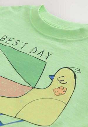 T-shirt vert clair avec un oiseau jaune de style cartoon et des formes géométriques, avec un texte noir indiquant "MEILLEURE JOURNÉE" au-dessus du design.