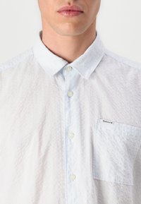 Camisa texturizada azul clara com colarinho abotoado, bolso frontal e um padrão subtil. Apresenta um tecido claro em contraste.