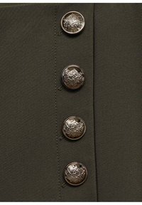 Quatre boutons métalliques avec un design en relief de blason complexe sur un panneau de tissu vert olive foncé avec des coutures visibles.