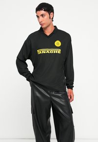 Schwarzes Langarmshirt mit V-Ausschnitt, das ein auffälliges gelbes Logo und den Text "ANACHE" zeigt. Getragen mit schwarzen Lederhosen.