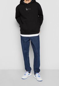 Sudadera negra con capucha y bolsillo canguro, detalle del logo en blanco, combinada con jeans de mezclilla azul y zapatillas blancas con acentos azules.