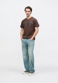 Καφέ βαμβακερό T-shirt με το κείμενο «PROBLEM OF DETAIL» σε μαύρο, σε συνδυασμό με ανοιχτό μπλε τζιν παντελόνι και πράσινα αθλητικά. Κανονική εφαρμογή και στυλ casual.