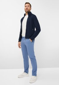 Marine blauwe zip-up jas over een wit T-shirt, gecombineerd met lichtblauwe broeken en witte sneakers. De jas heeft een hoge kraag en ribgebreide manchetten.