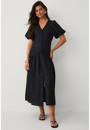 Kvinde iført en sort midikjole med pufærmer, knapper foran, frontslids, guldbøjle og sorte sandaler står mod en hvid baggrund.