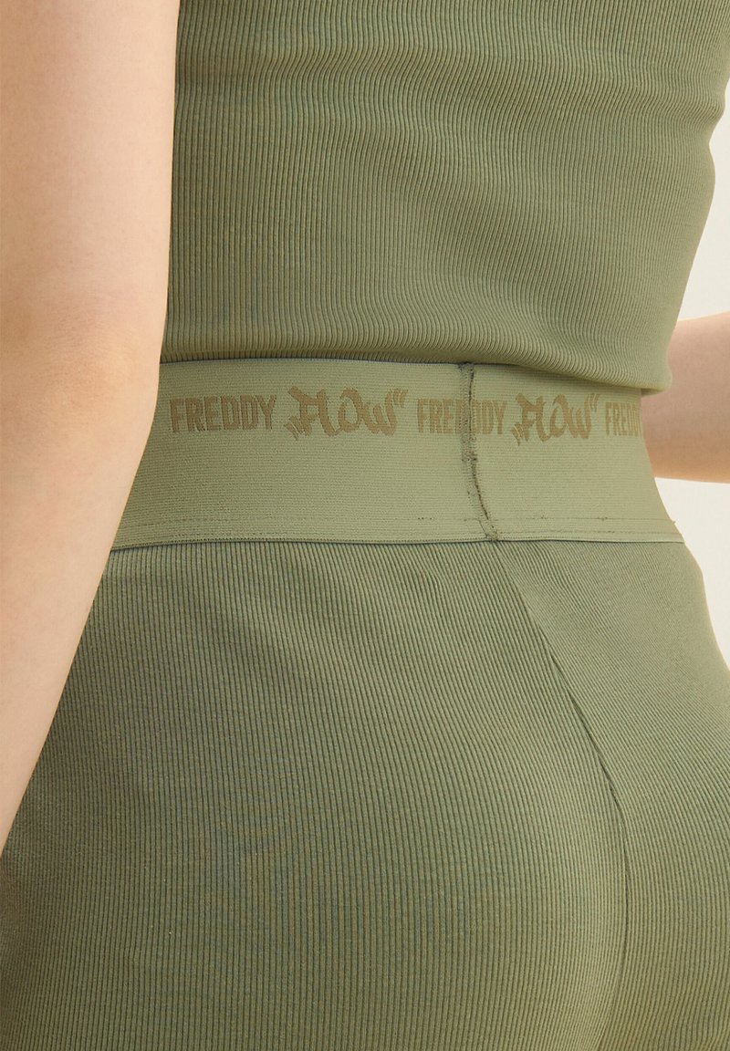 Gros plan sur une personne portant un haut assorti en maille côtelée vert olive et un legging taille haute avec l'inscription "FREDDY FLOW" sur la ceinture.