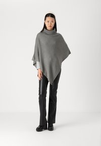 Grå stickad poncho med hög krage, som har en lös och flödande silhuett och texturerad tyg, i kombination med svarta fusk läderbyxor.