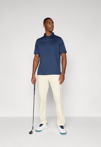 Marineblå polo med kortærmer, blødt stof og krave. Parret med lyse beige bukser og hvide golf sko med marineblå detaljer.
