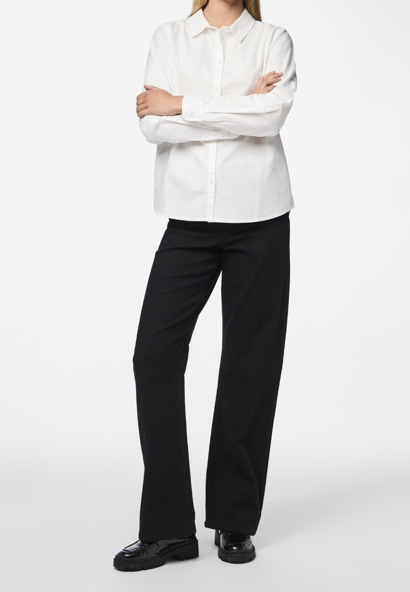 Femme debout, bras croisés, portant une chemise blanche boutonnée, un pantalon noir à jambes larges et des chaussures noires sur fond blanc.