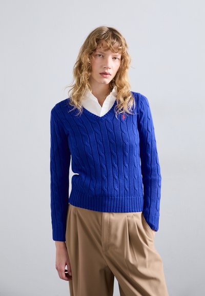 Jersey de punto azul con cuello en V y puños y dobladillo acanalados, que presenta un pequeño logo rojo en el pecho, combinado con pantalones beige.