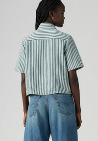 Camicia corta a maniche corte di colore azzurro chiaro con righe bianche verticali. Presenta un colletto classico e un delicato dettaglio cucito sulle cuciture.