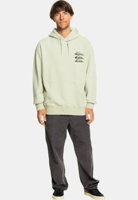 Sudadera verde claro con un bolsillo frontal y tres diseños gráficos impresos en el lado izquierdo del pecho; combinada con pantalones de pana gris oscuro y zapatos negros.