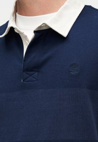 Άνδρας που φορεί μπλε σκούρο πόλο πουκάμισο με λευκό γιακά και μικρό κεντημένο λογότυπο δέντρου στο στήθος.