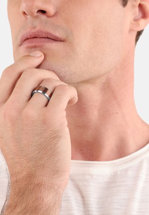 Metallring mit einem gebürsteten Kupferband und polierten silbernen Akzenten. Glatte Oberfläche, am Finger getragen, während eine Hand das Kinn berührt.