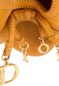 Gros plan d'un sac en cuir beige avec des surpiqûres blanches et des lettres métalliques dorées suspendues formant le mot « Dior », attachées à des lanières en cuir.