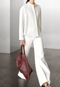 Chemise à carreaux blancs, pantalon blanc ample et sac fourre-tout tissé rouge texturé. La chemise a un col montant et des manches longues.