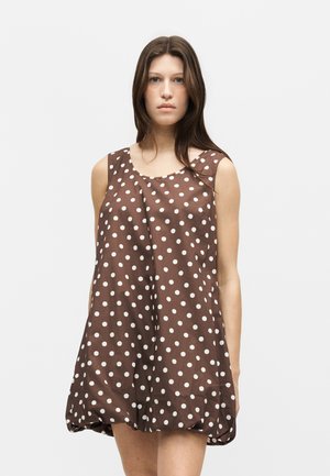 Femme portant une robe marron sans manches à pois blancs, debout devant un fond clair uni, regardant devant avec une expression neutre.