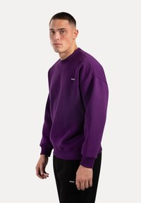 Sudadera morada con cuello redondo y puños acanalados. Con un pequeño logo en el pecho. Textura de tela suave. Combinada con pantalones negros.