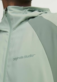 Svetlo zelena zip-up jakna z gladko teksturo, kontrastnimi paneli in logotipom "Originals Studio" na levem prsnem predelu.