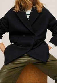 Tummansininen kaksirivinen takki kalanruotokuvioisella pinnalla, suuret lovelliset kaulukset ja ylisuurilla hihoilla. Päällä oli oliivinvihreät housut.