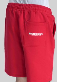 Červené bavlněné šortky s elastickým pasem, s velkou náprsní kapsou a bílým textem "MULTIPLY" na zadní straně. Hladká textilie.