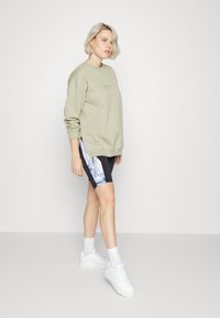 Sweatshirt verde-oliva com punhos canelados, combinado com calções de bicicleta pretos com um padrão de mármore azul. Ténis brancos e meias até ao tornozelo completam o visual.